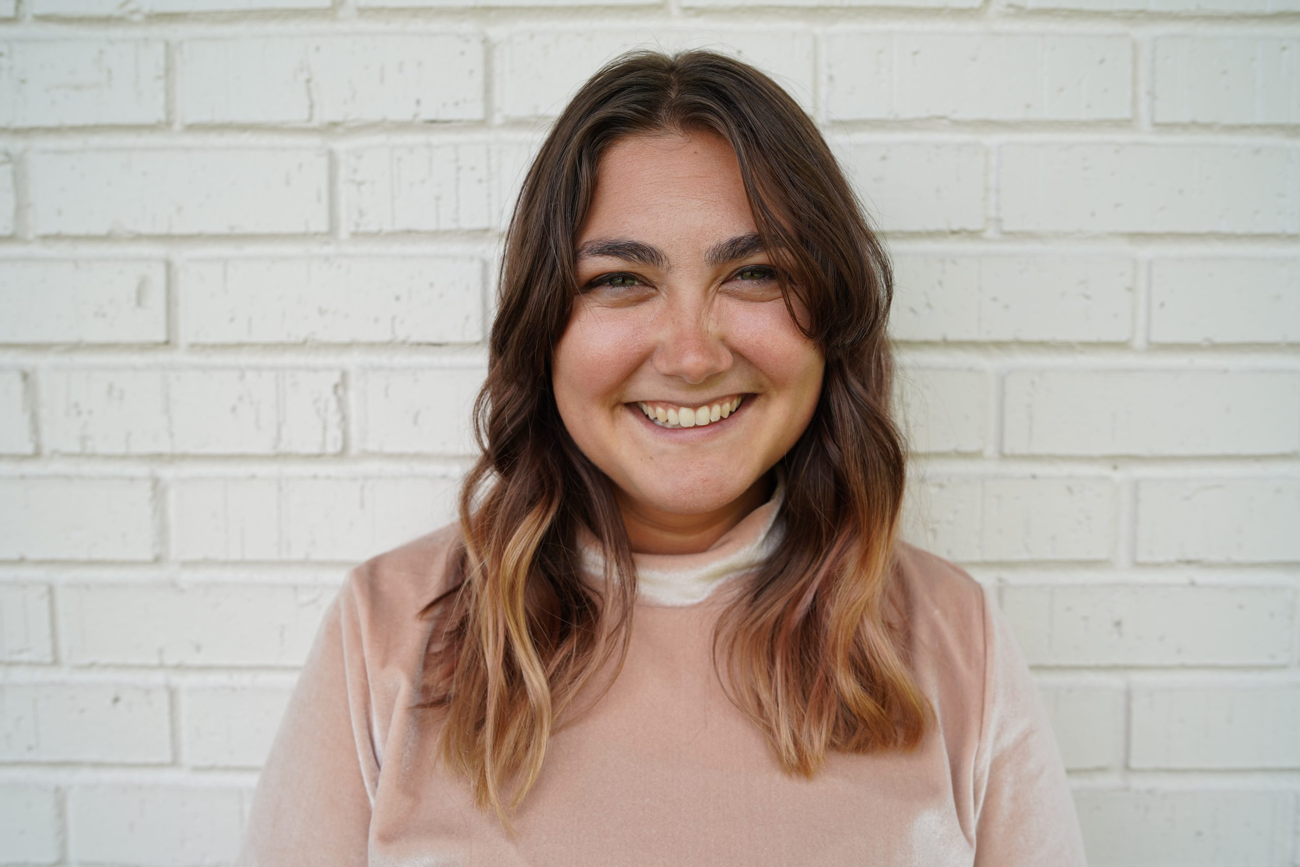 woman with brown and blond hair smiling in front of a white brick wall. She has on a dusty pink turtleneck shirt.